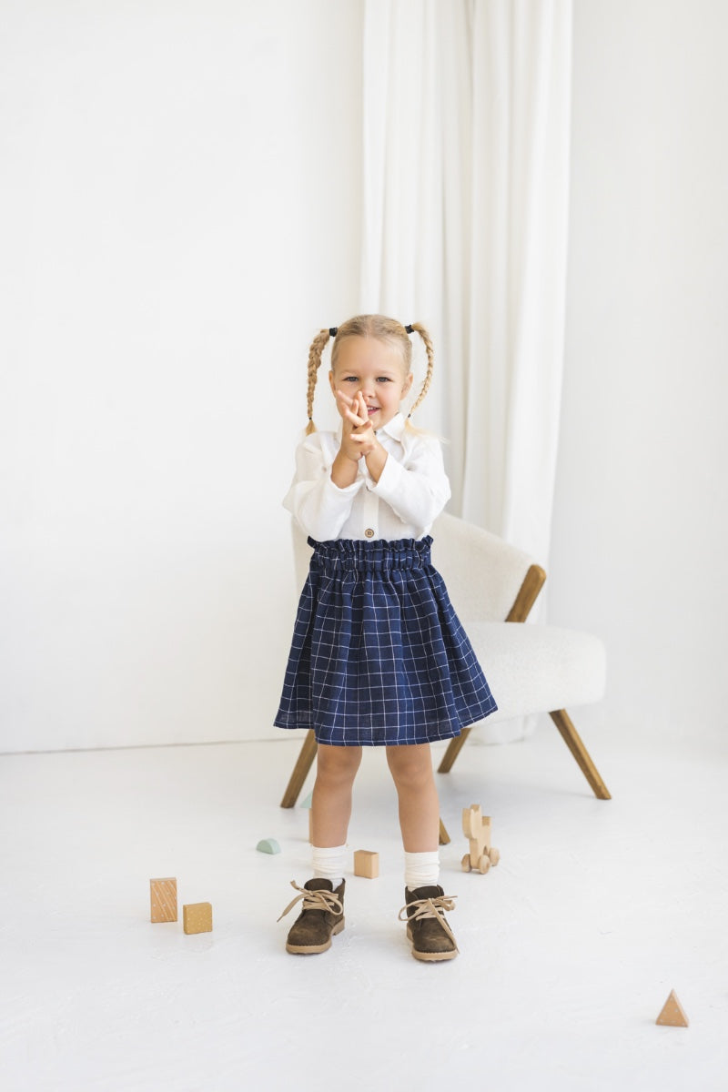 Young girl in a white blouse and blue plaid linen skirt standing in a minimalistic room.