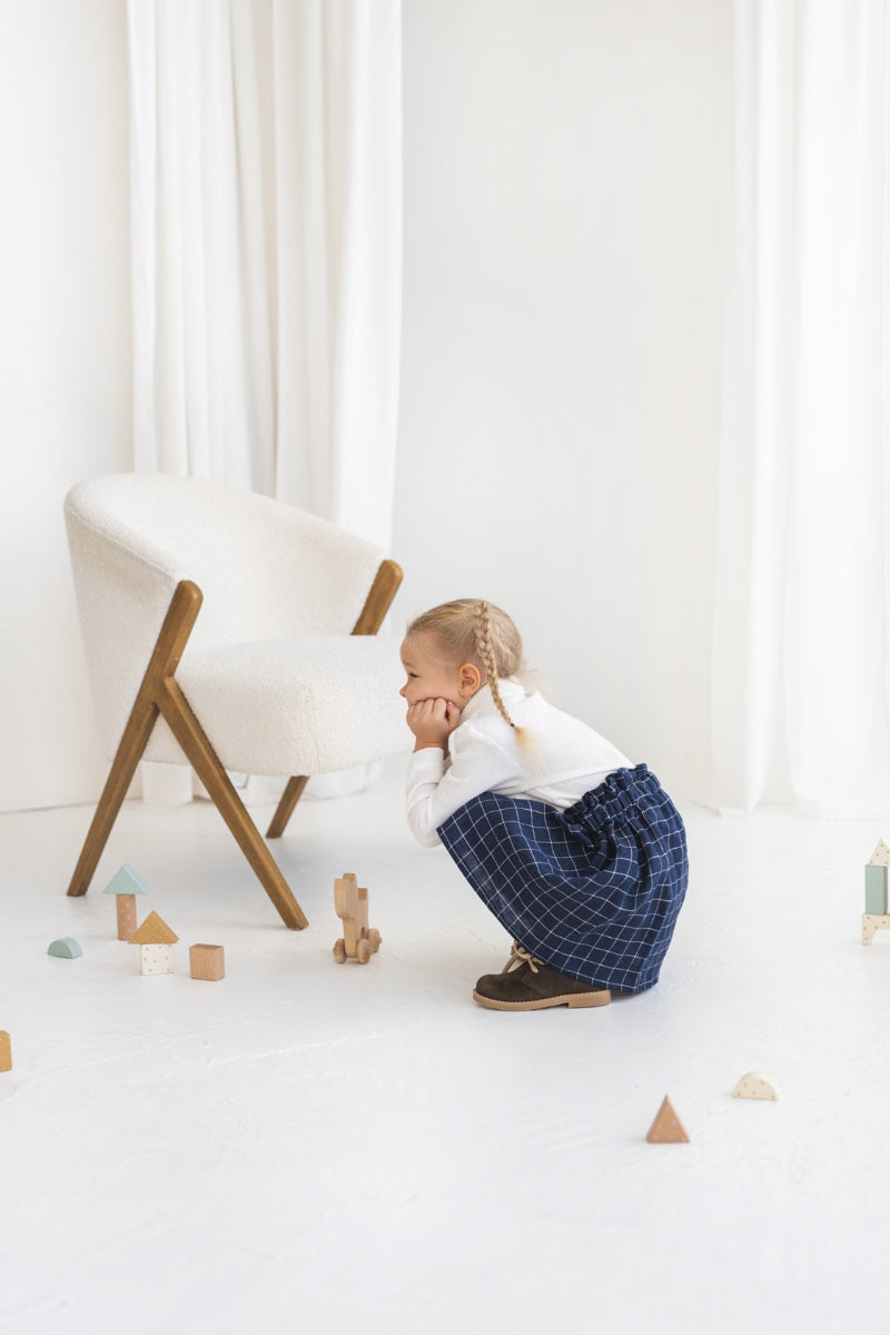 Young girl in a white blouse and blue plaid linen skirt standing in a minimalistic room.
