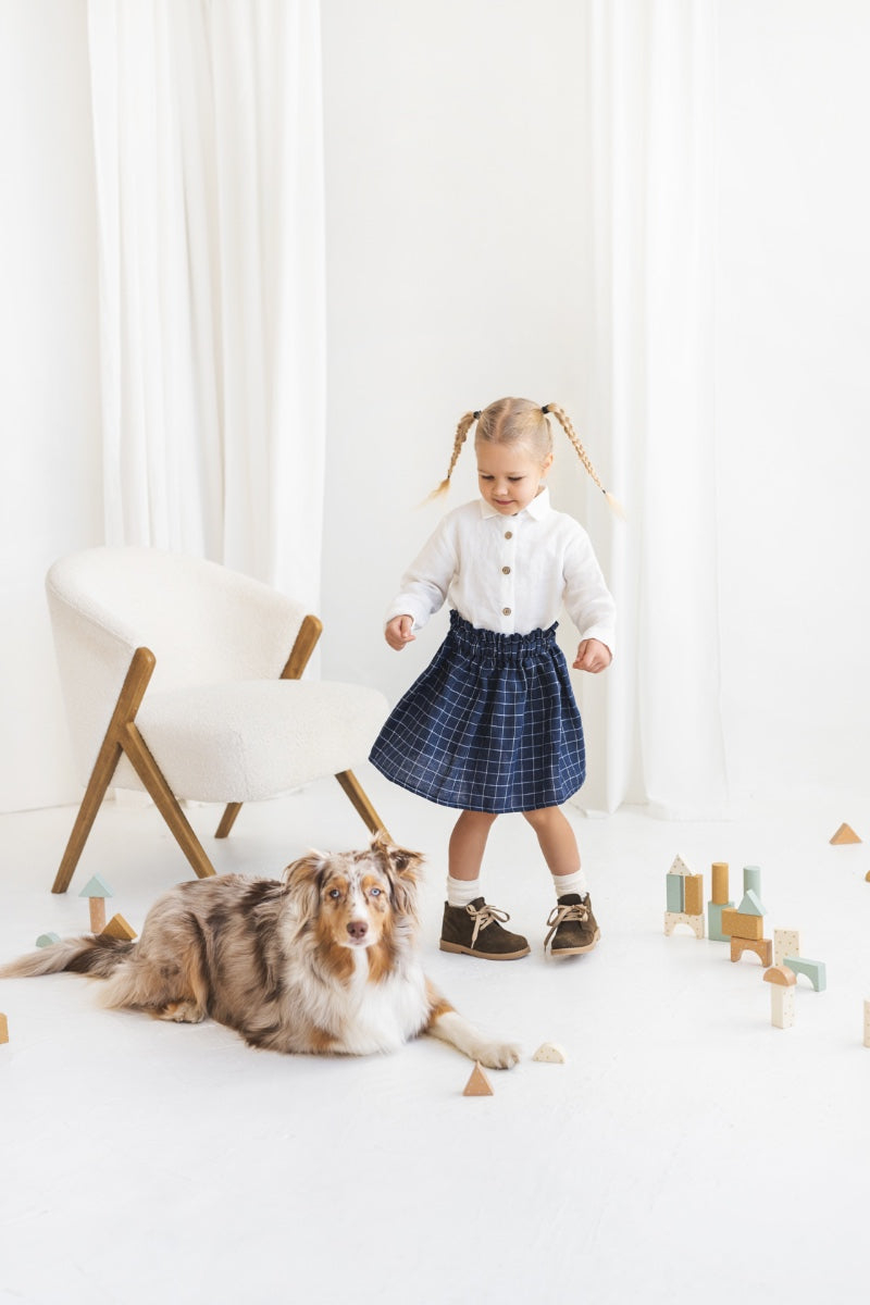 Young girl in a white blouse and blue plaid linen skirt standing in a minimalistic room.