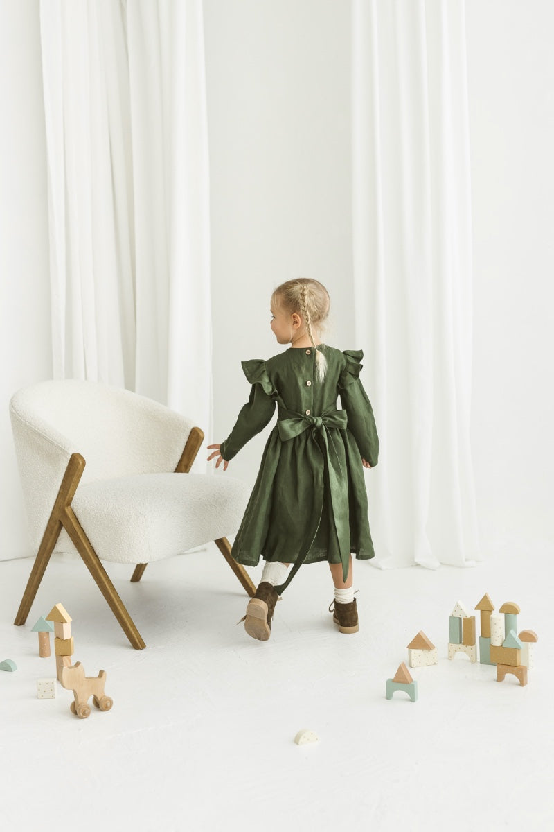 Child in a green linendress standing near a white chair with wooden legs on a white floor.