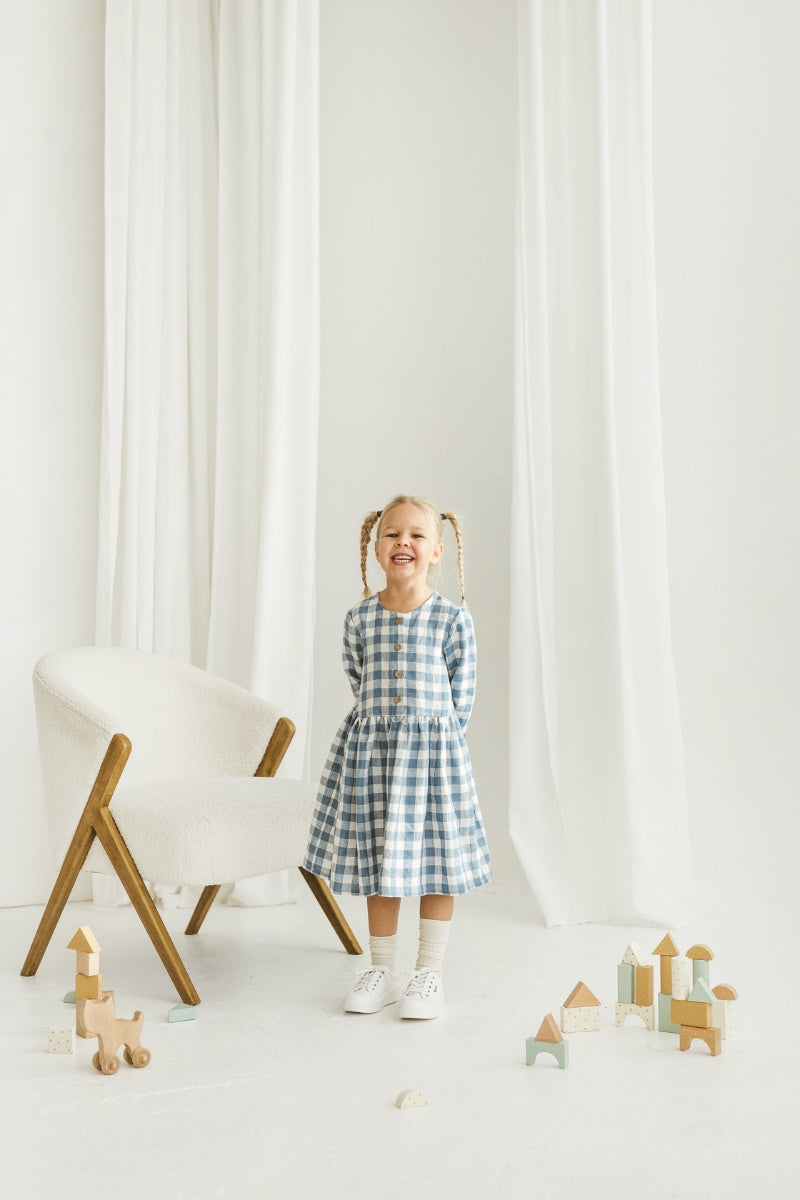 Girl in a blue checkered linen dress standing in a room with white curtains and wooden toys on the floor.