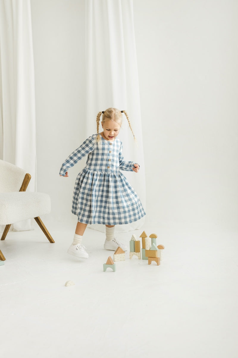 Child in a blue checkered linen dress standing in a minimalistic room with white curtains and wooden toys.