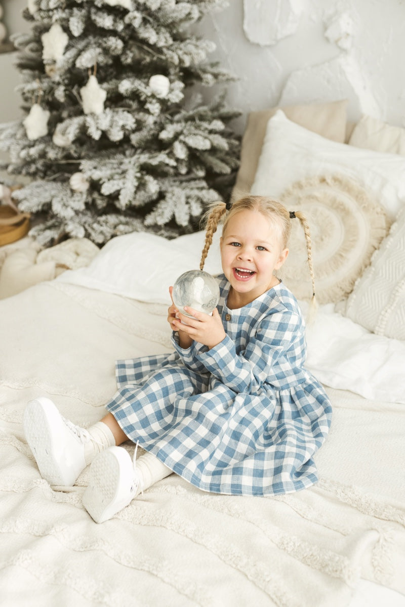 Child in a blue checkered linen dress holding a silver ornament in front of a decorated Christmas tree.