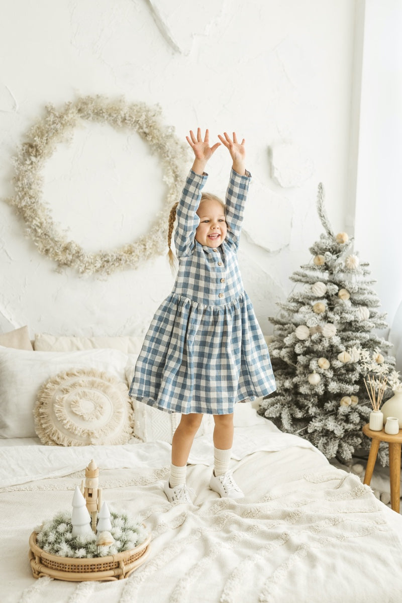 Girl in a linen gingham dress standing in a room with a Christmas tree and wreath.