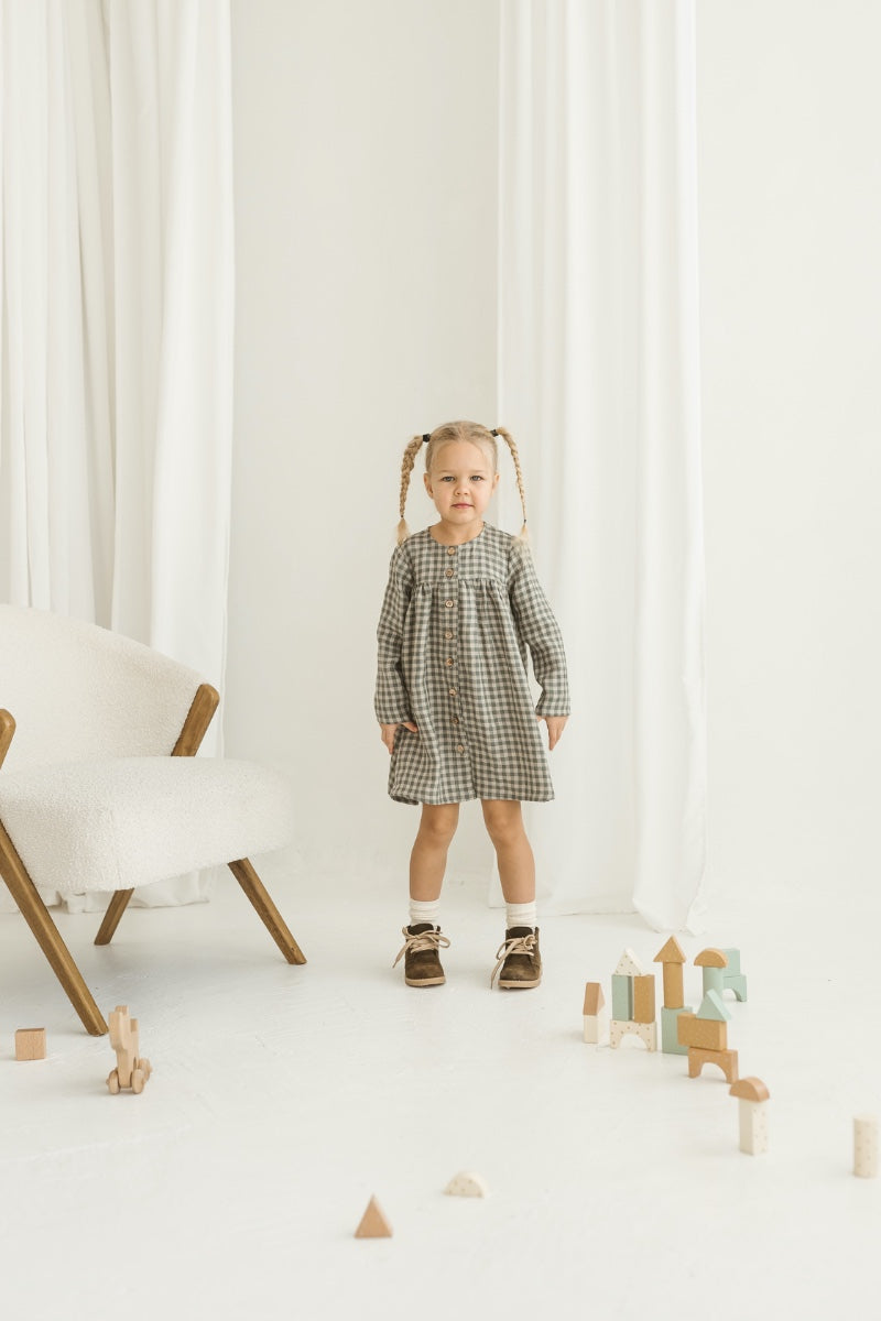 Young girl in a grey linen checkered dress standing in a minimalistic room with white curtains and wooden toys.