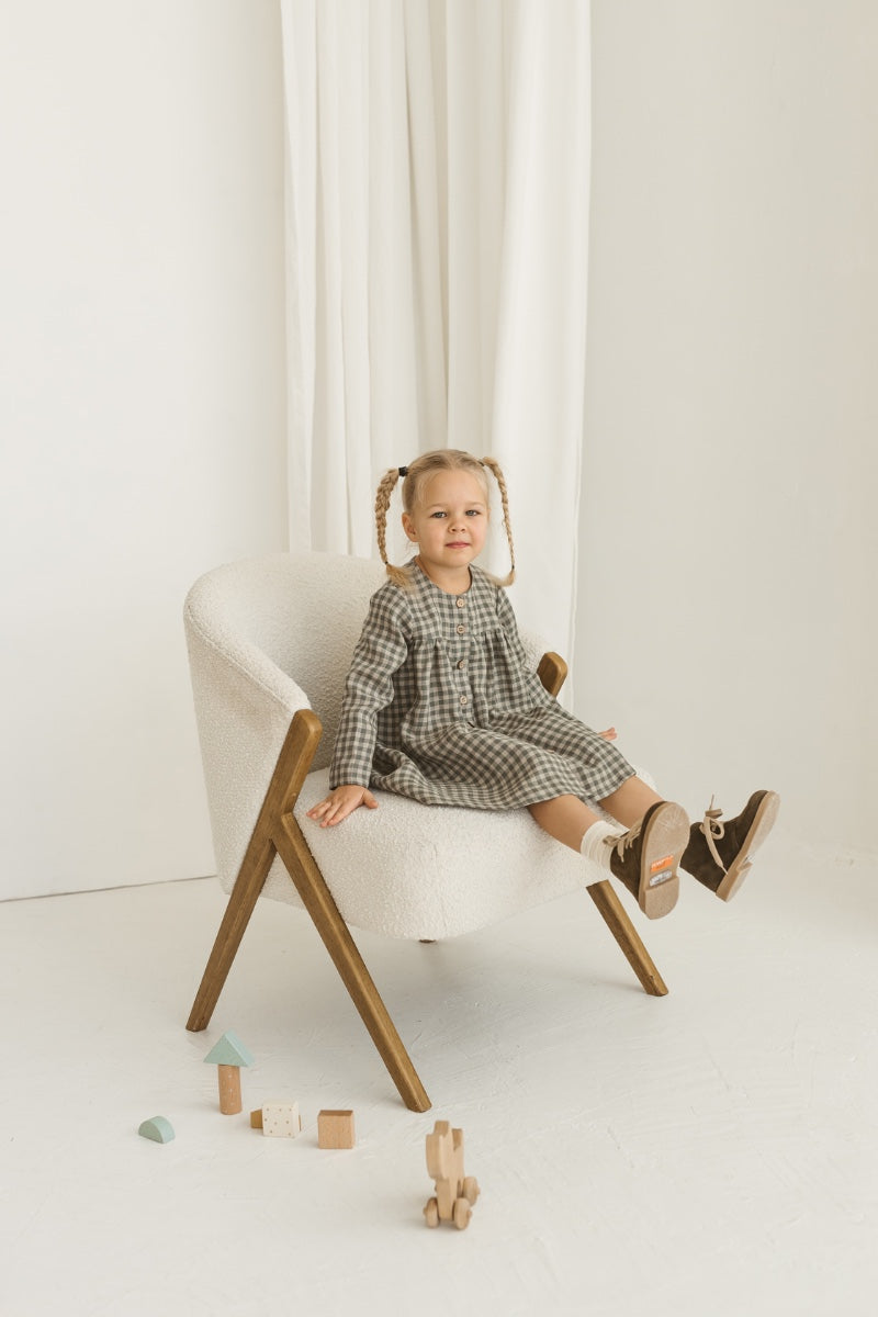 Young girl in a grey linen checkered dress standing in a minimalistic room with white curtains and wooden toys.