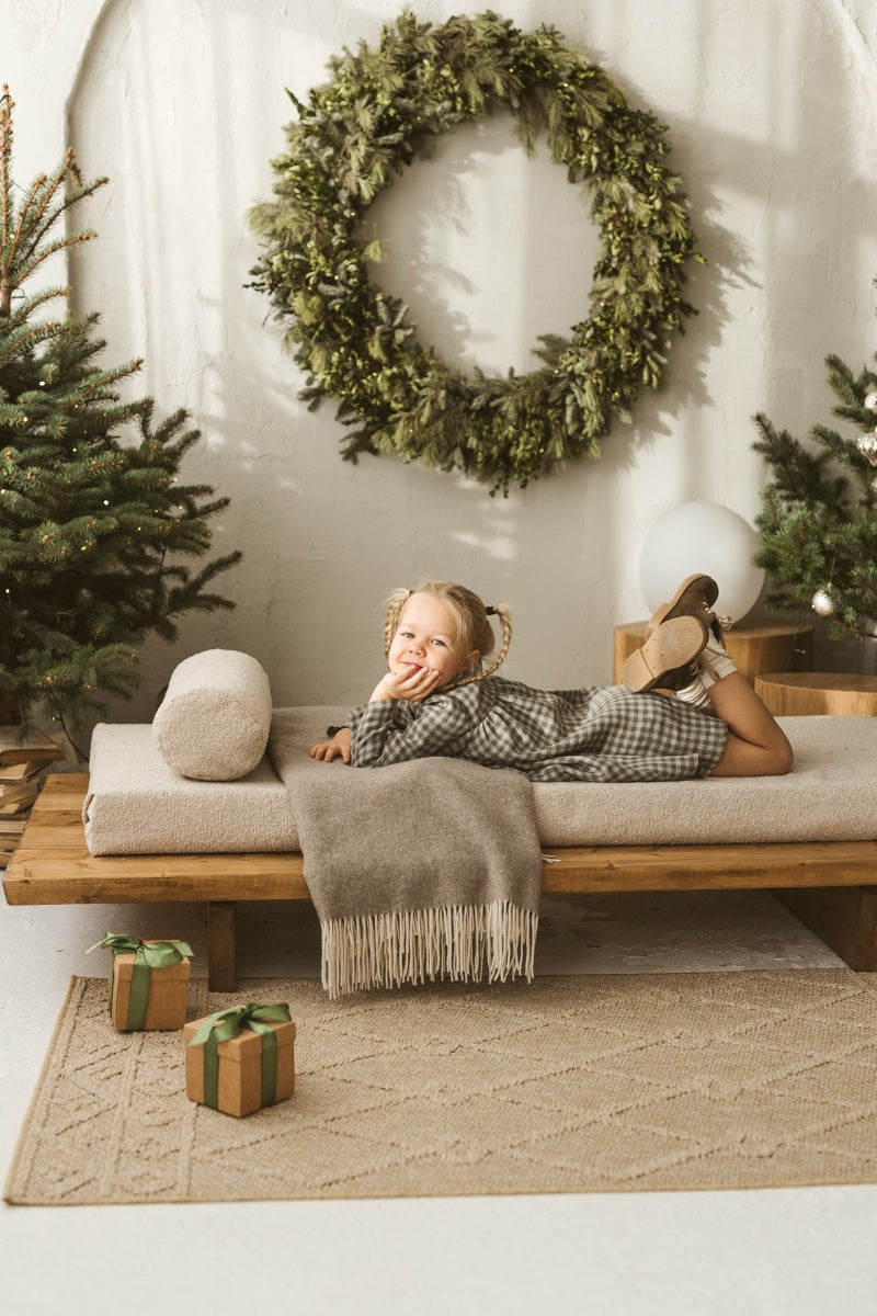 Young girl in a grey linen checkered dress standing in a minimalistic room with white curtains and wooden toys.
