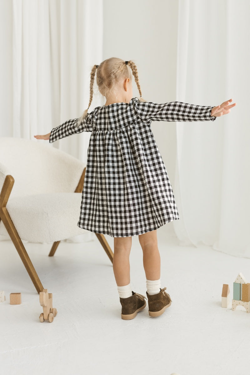 Child in a checkered linen dress standing on a white floor with wooden toys.