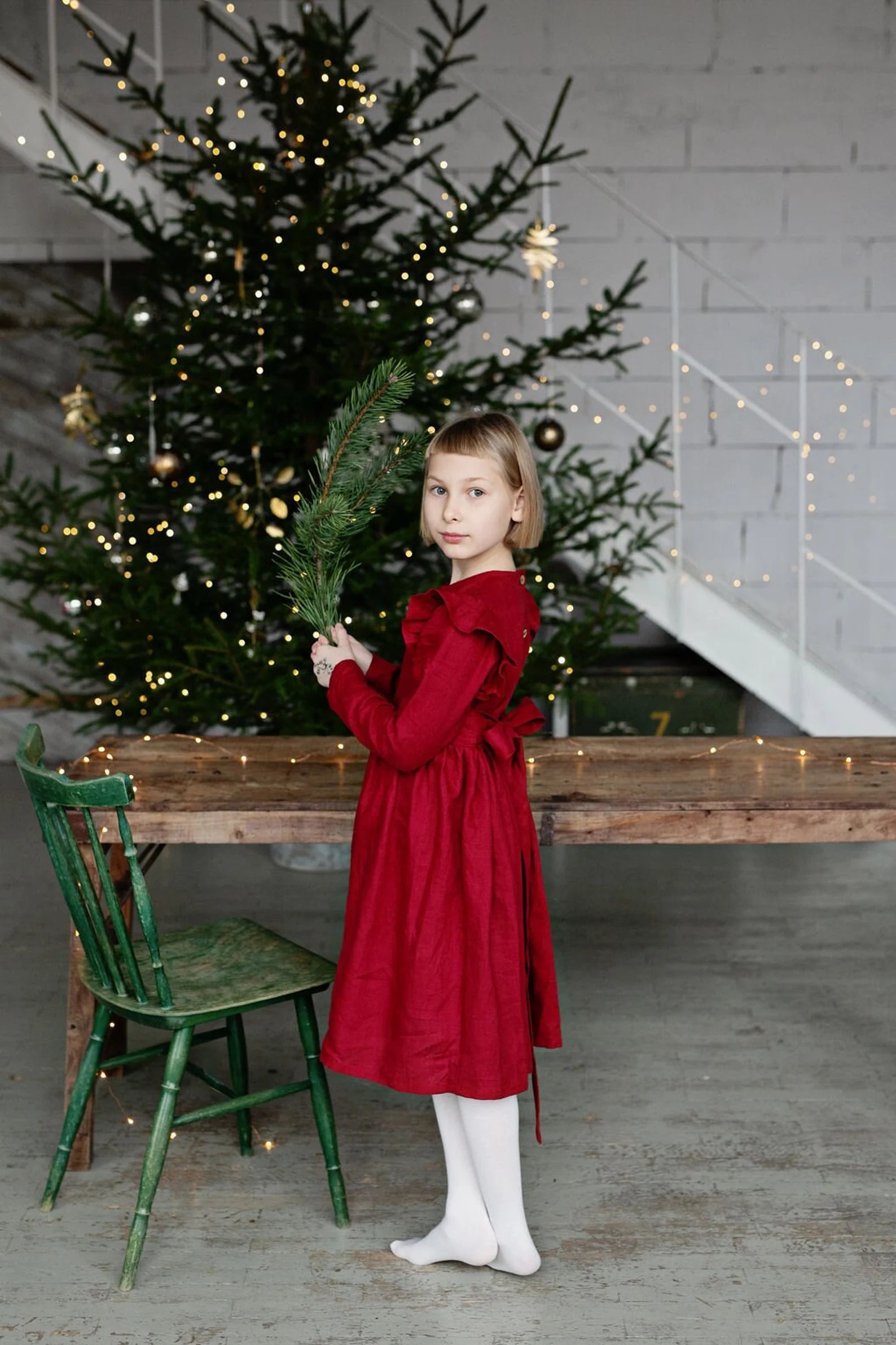 Young girl in a linen red dress holding a Christmas tree branch in front of a decorated Christmas tree.