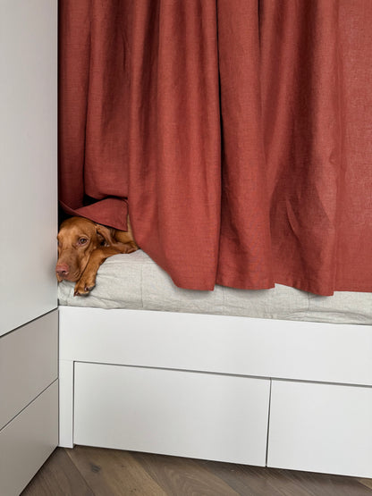Dog peeking out from behind a red curtain in a room with white walls and wooden floor.

