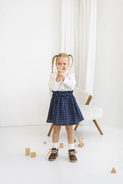 Young girl in a white blouse and blue plaid linen skirt standing in a minimalistic room.


