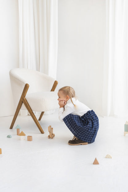 Young girl in a white blouse and blue plaid linen skirt standing in a minimalistic room.
