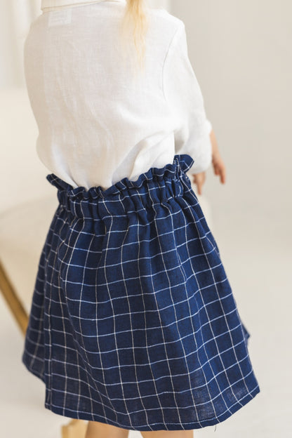 Young girl in a white blouse and blue plaid linen skirt standing in a minimalistic room.
