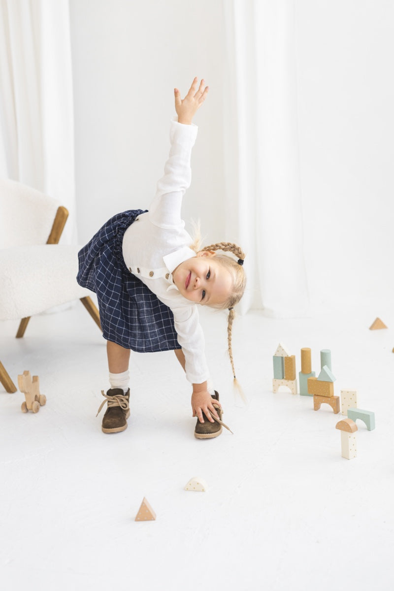 Young girl in a white blouse and blue plaid linen skirt standing in a minimalistic room.
