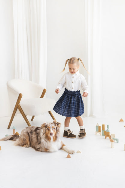 Young girl in a white blouse and blue plaid linen skirt standing in a minimalistic room.
