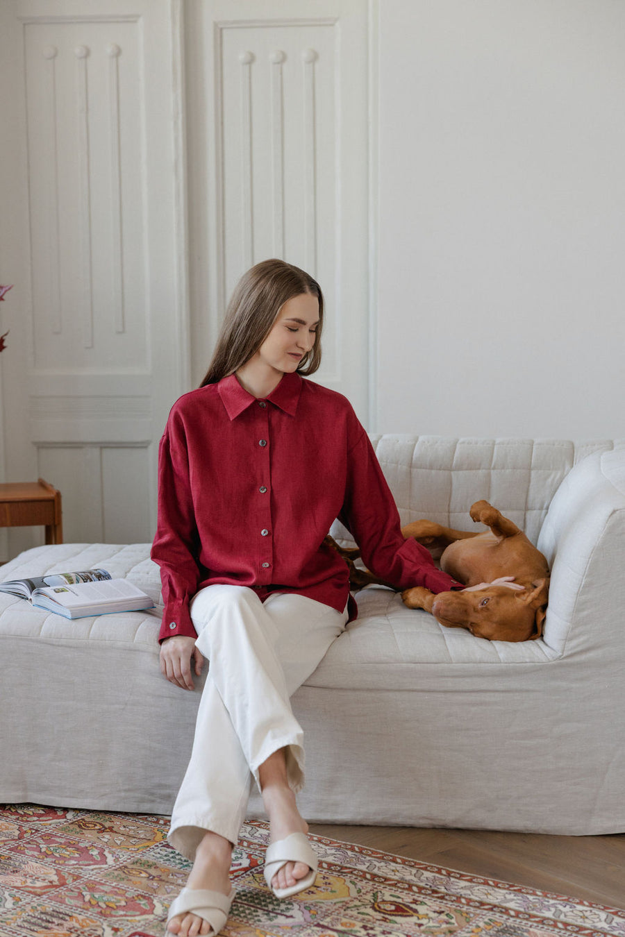 Woman in a red shirt sitting on a white couch with a dog, in a room with a patterned rug and wooden side table.