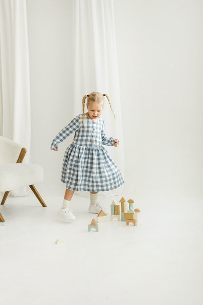 Child in a blue checkered linen dress standing in a minimalistic room with white curtains and wooden toys.

