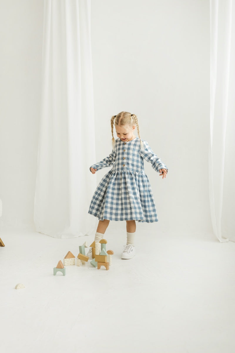 Girl in a blue checkered linen dress standing in a room with white curtains and wooden toys on the floor.
