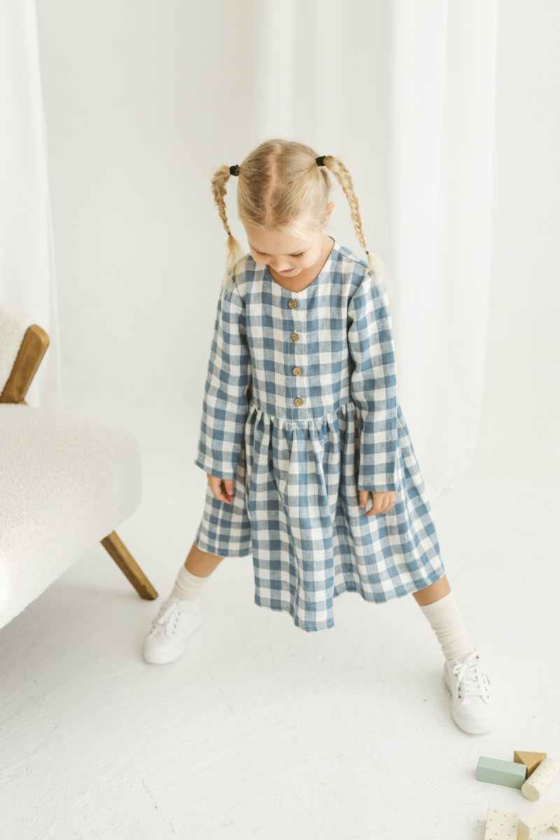 Girl in a blue checkered linen dress standing in a room with white curtains and wooden toys on the floor.
