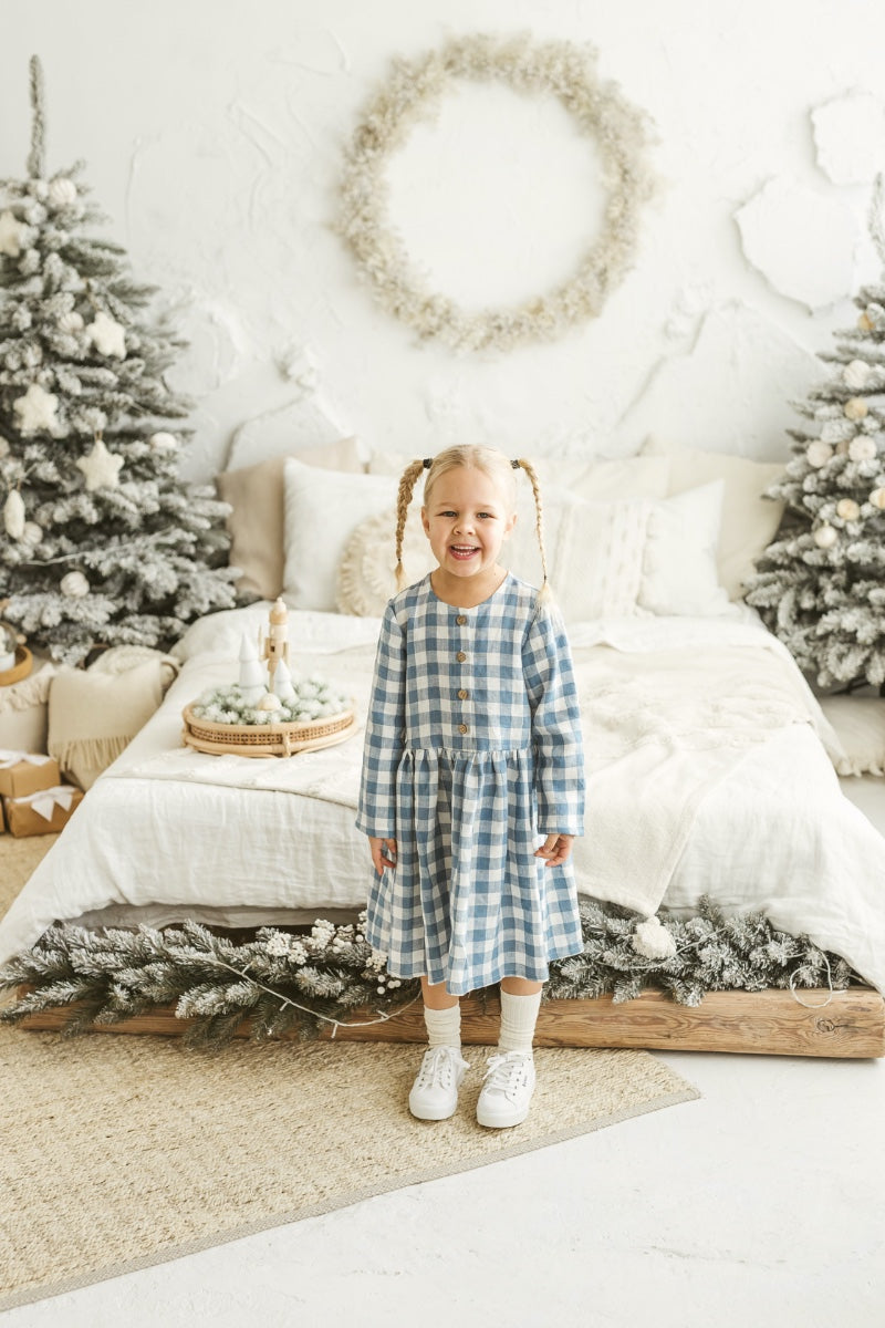 Girl in a blue checkered linen dress standing in a decorated room with Christmas trees and wreaths.

