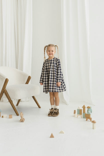 Young girl in a checkered linen dress standing in a minimalistic room with white curtains and wooden toys on the floor.


