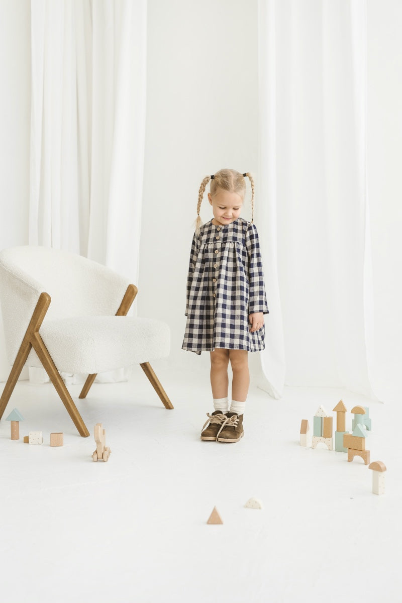 Young girl in a checkered linen dress standing in a minimalistic room with white curtains and wooden toys on the floor.
