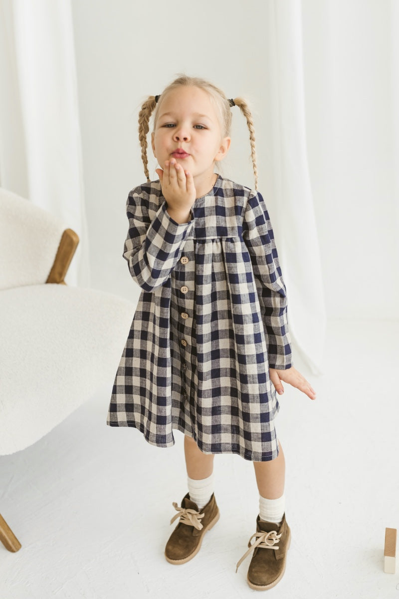 Young girl in a checkered linen dress standing in a minimalistic room with white curtains and wooden toys on the floor.
