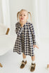 Young girl in a checkered linen dress standing in a minimalistic room with white curtains and wooden toys on the floor.
