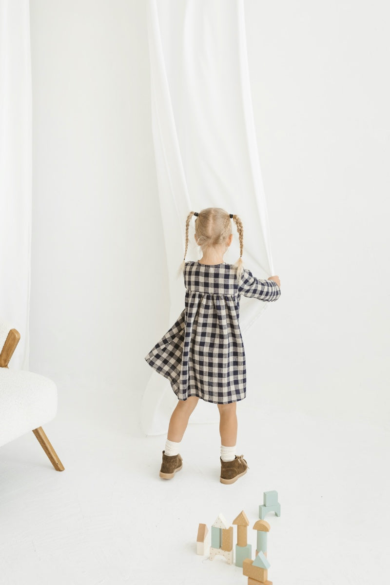 Young girl in a checkered linen dress standing in a minimalistic room with white curtains and wooden toys on the floor.
