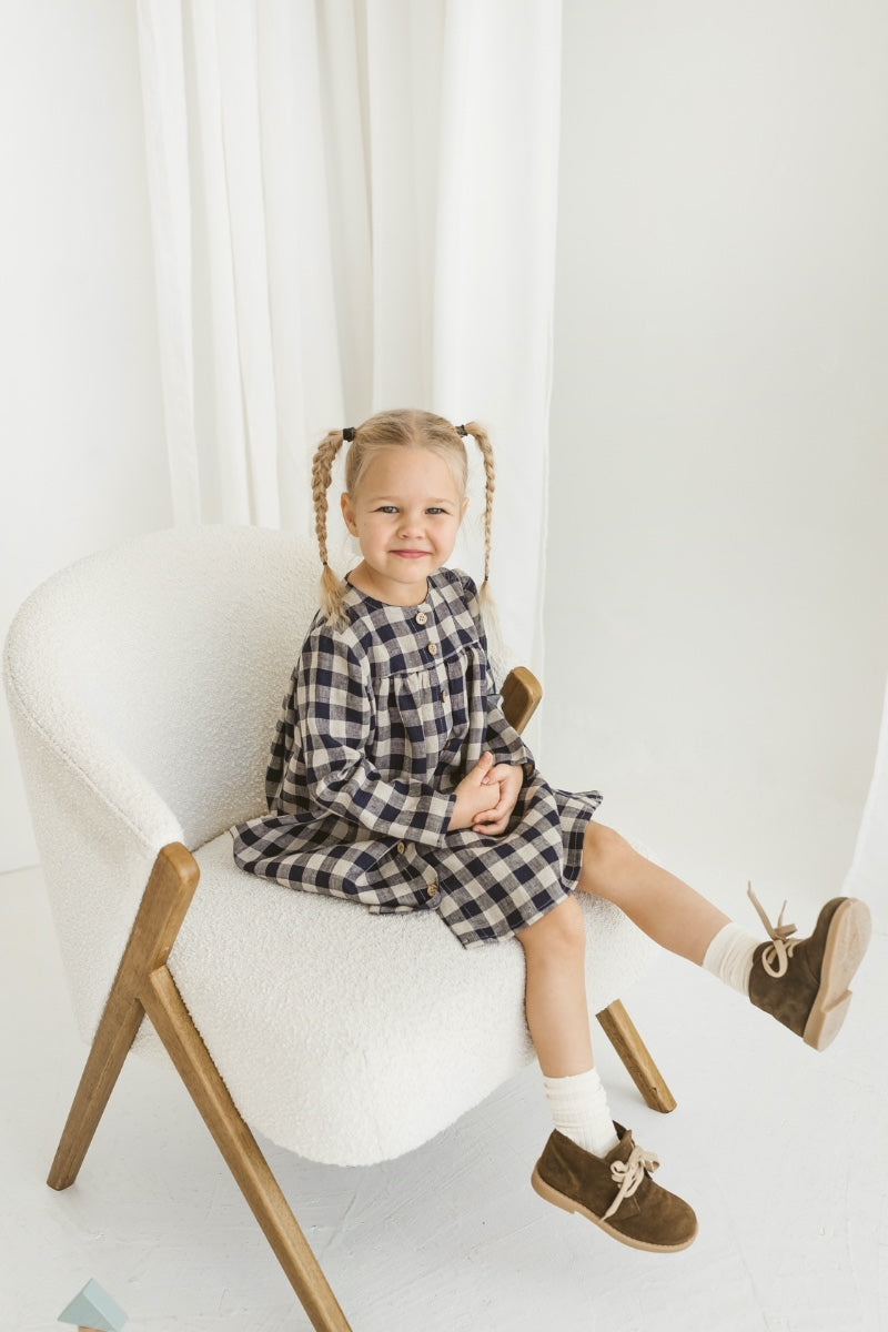 Young girl in a checkered linen dress standing in a minimalistic room with white curtains and wooden toys on the floor.
