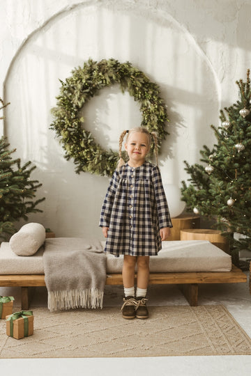 Young girl in a checkered linen dress standing in a minimalistic room with white curtains and wooden toys on the floor.
