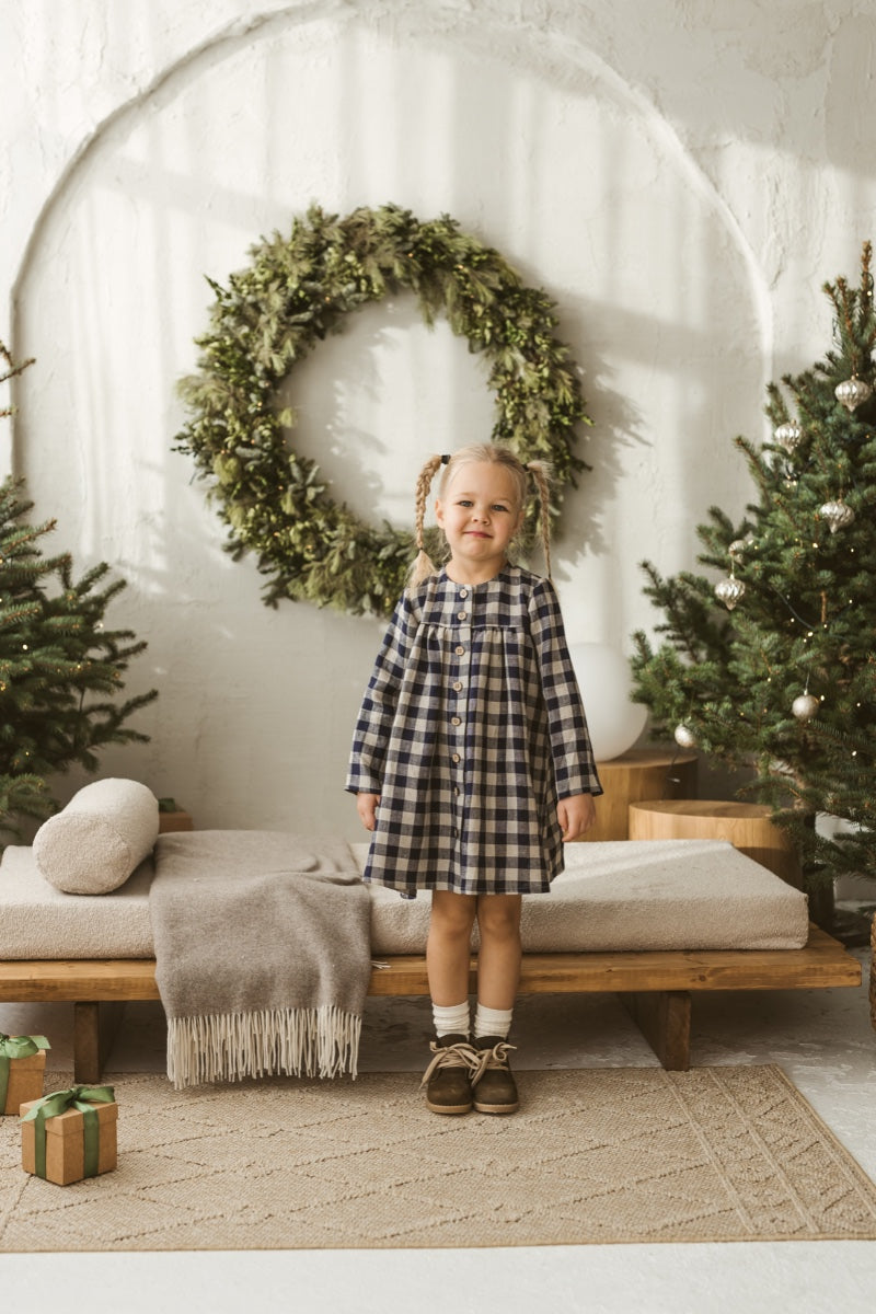 Young girl in a checkered linen dress standing in a minimalistic room with white curtains and wooden toys on the floor.
