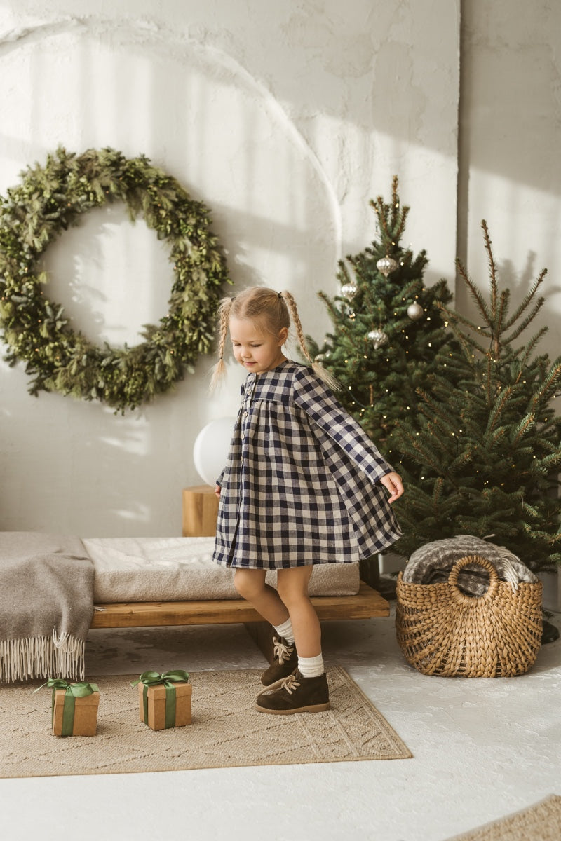 Young girl in a checkered linen dress standing in a minimalistic room with white curtains and wooden toys on the floor.
