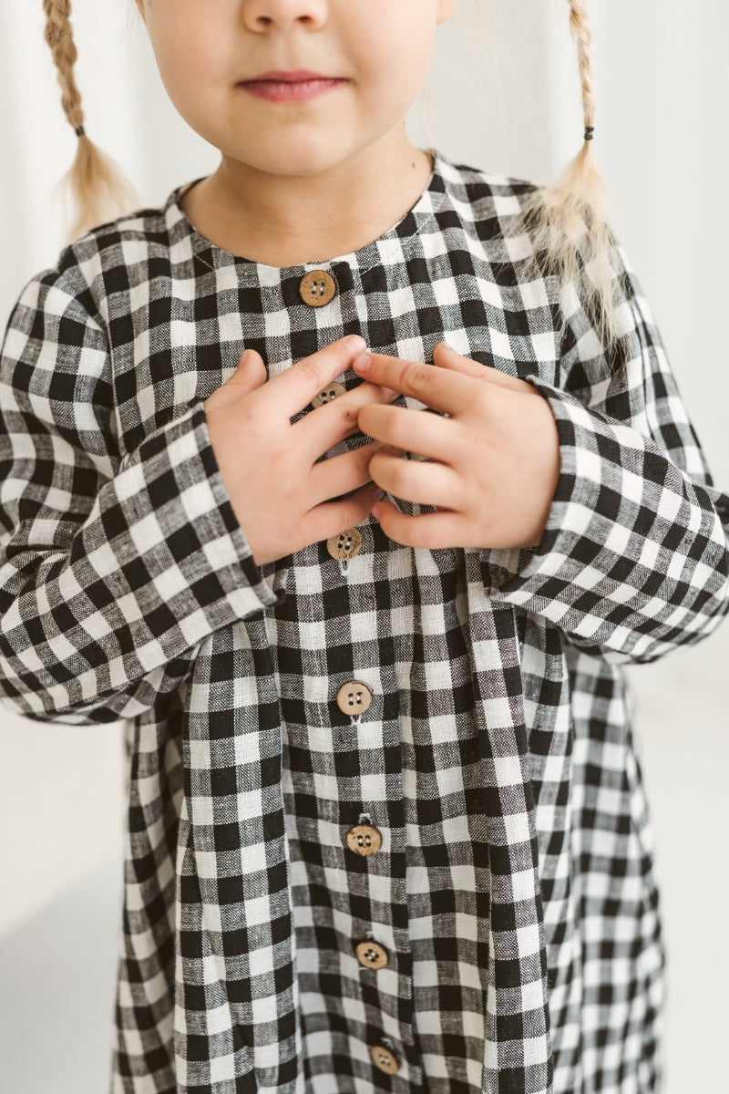 Child in a checkered linen dress standing on a white floor with wooden toys.
