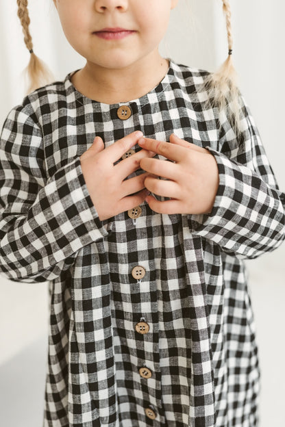 Child in a checkered linen dress standing on a white floor with wooden toys.
