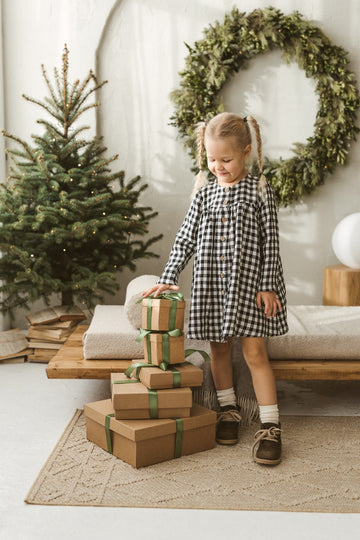 Child in a checkered linen dress standing on a white floor with wooden toys.
