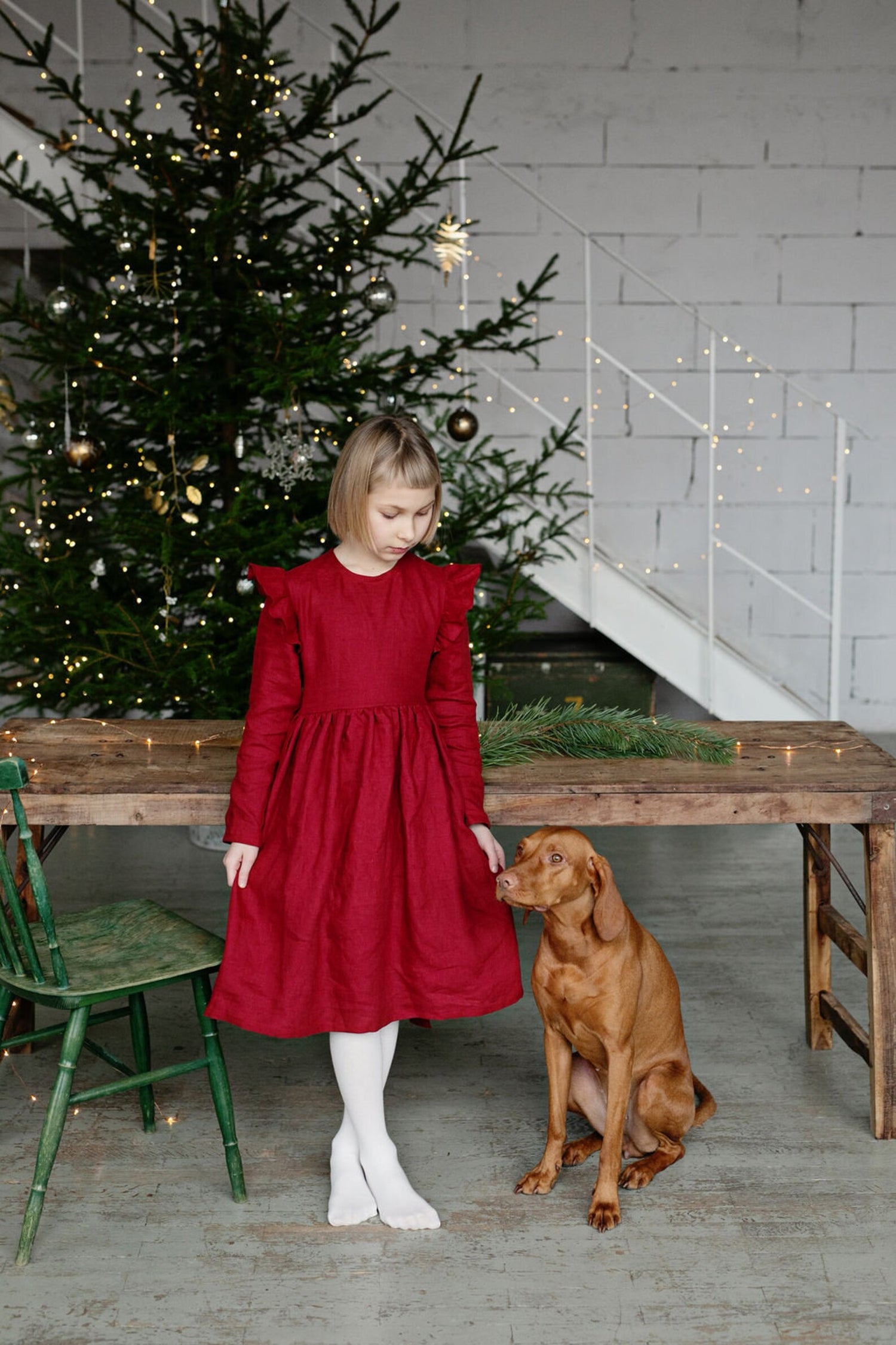 Young girl in a linen red dress standing next to a dog in a festive setting with a Christmas tree.