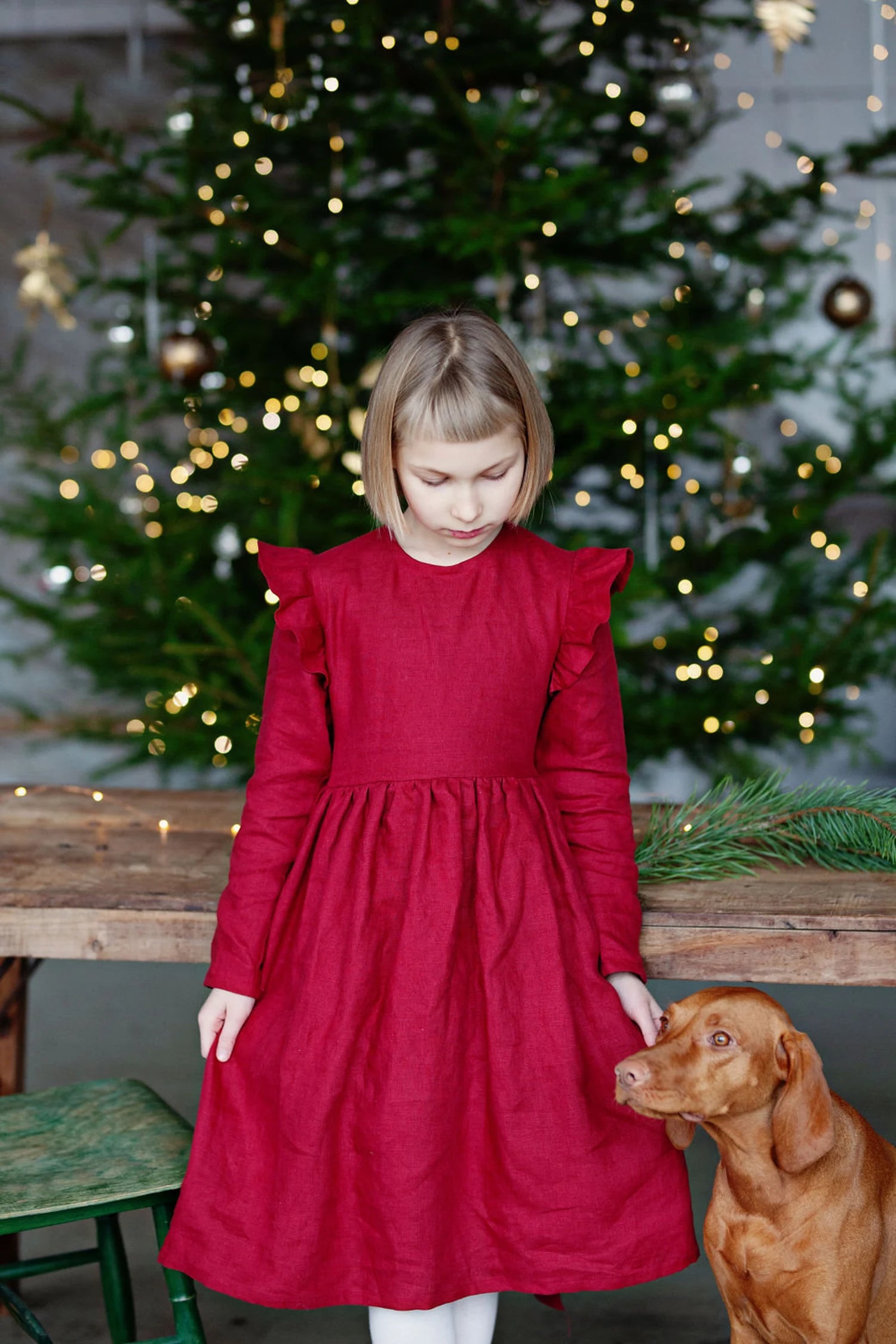 Girl in a red linen dress sitting in front of a decorated Christmas tree.
