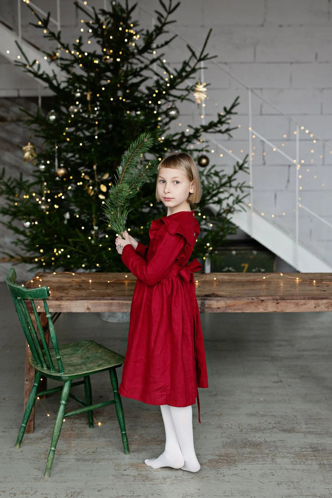 Young girl in a linen red dress holding a Christmas tree branch in front of a decorated Christmas tree.