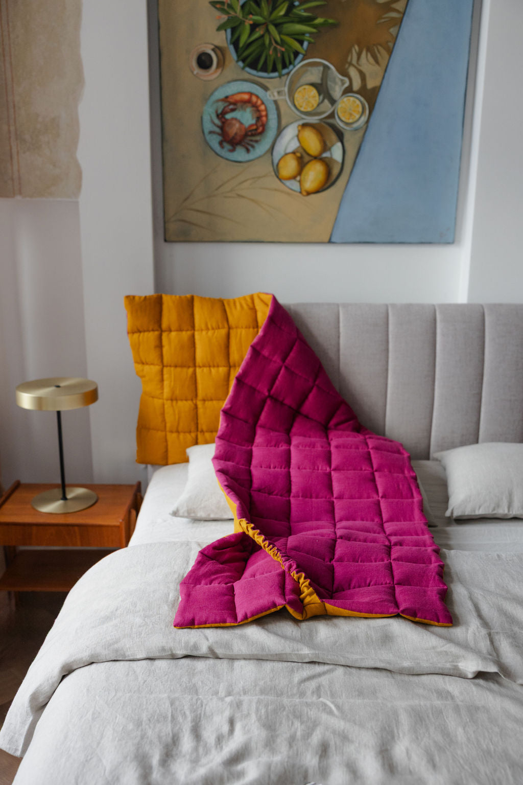 Pink and yellow quilt draped over a bed in a bedroom with a painting on the wall.