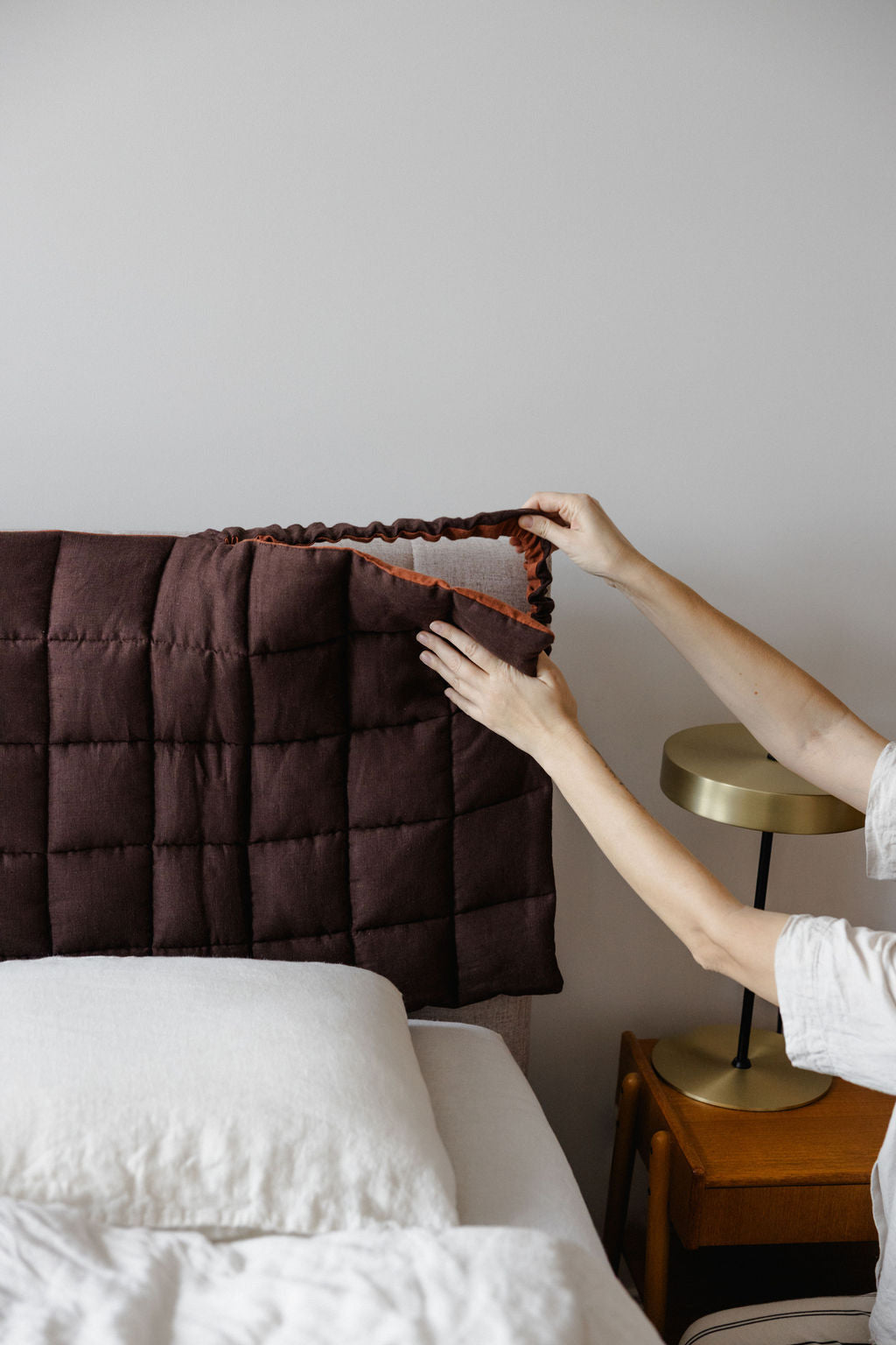 Person adjusting a brown quilt on a bed with a neutral background