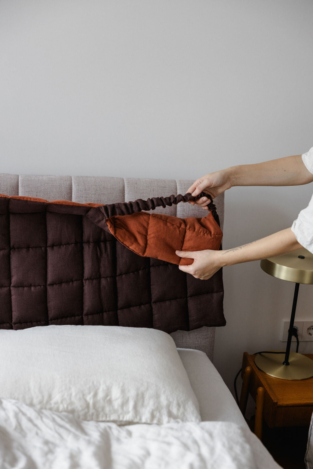 Person adjusting a brown pillow on a bed with a neutral background