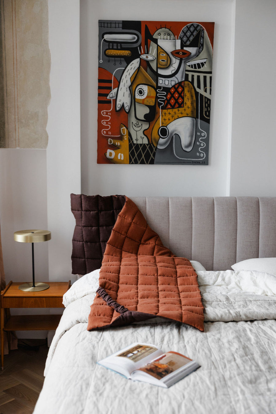 Bedroom with a bed featuring a quilt and pillows, a nightstand with a lamp, and a colorful abstract painting on the wall.