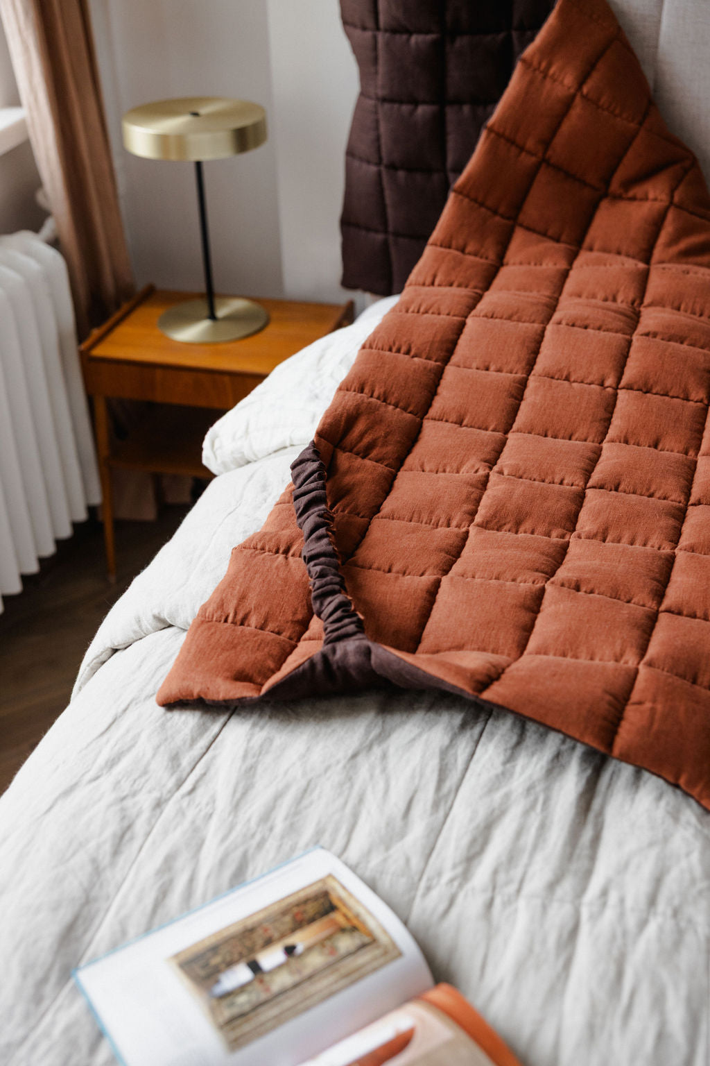 Brown quilted blanket draped over a bed with a side table and lamp in the background.