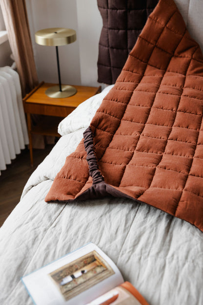 Brown quilted blanket draped over a bed with a side table and lamp in the background.