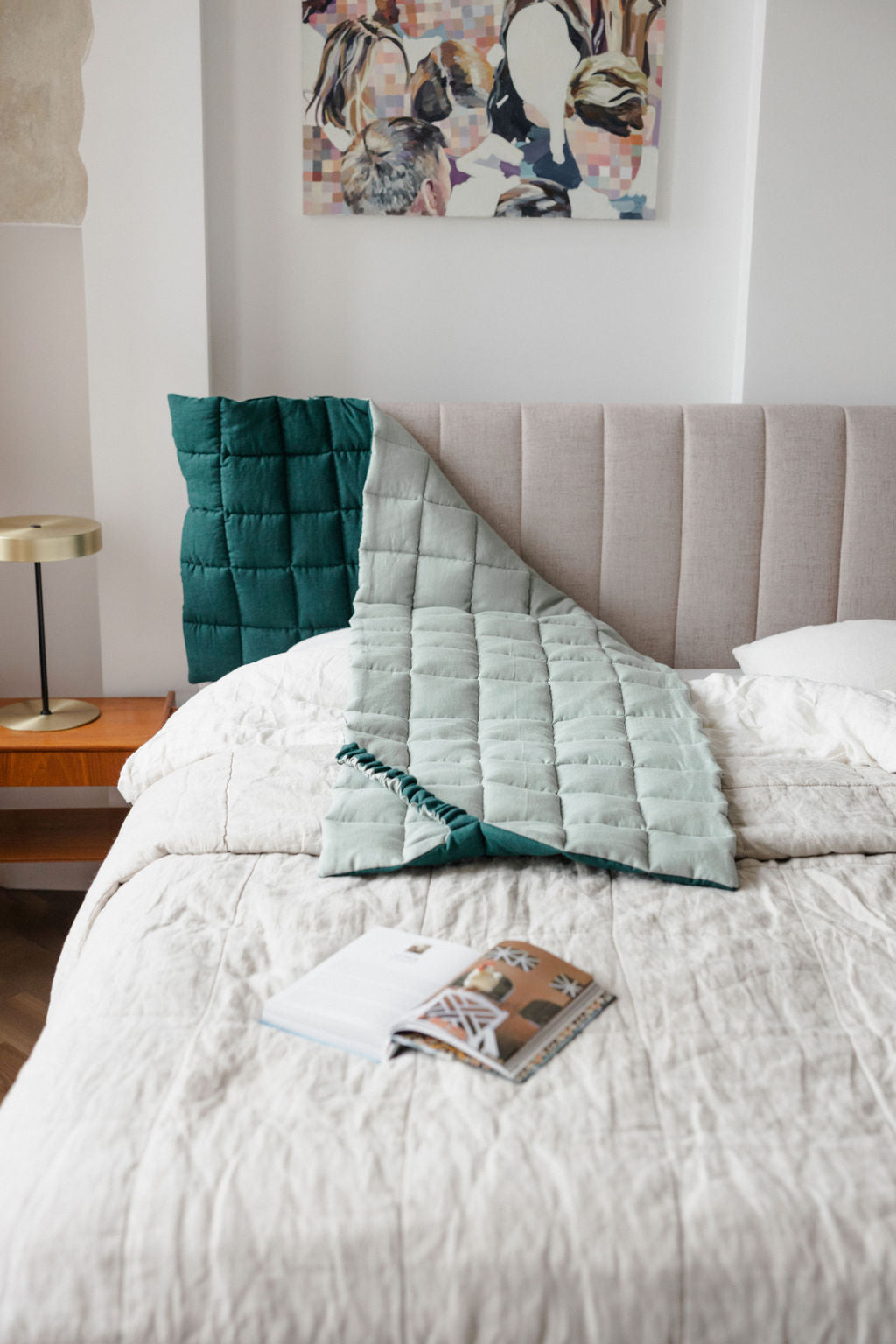 Bedroom with a bed featuring green and gray pillows, a magazine, and a lamp on a nightstand.