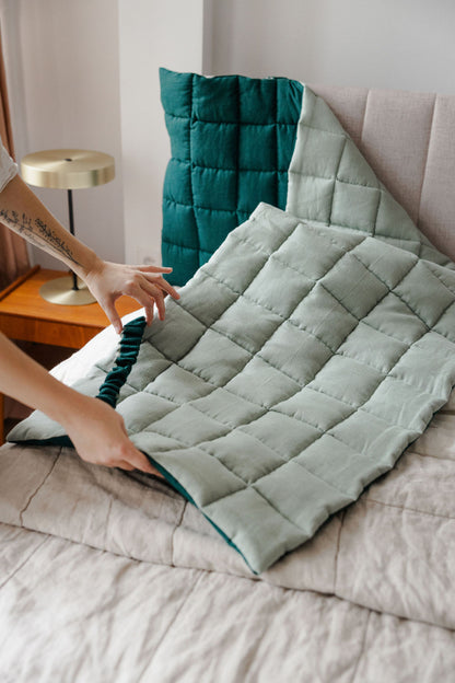 Person arranging a green quilt on a bed with a teal pillow in the background.