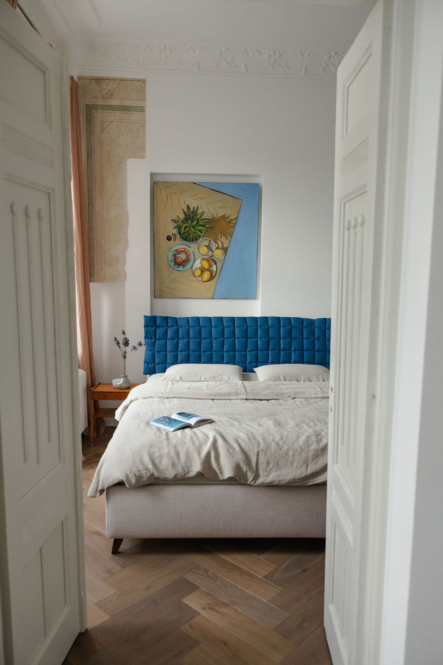 Bedroom with a blue tufted headboard and white bedding, featuring a painting on the wall.