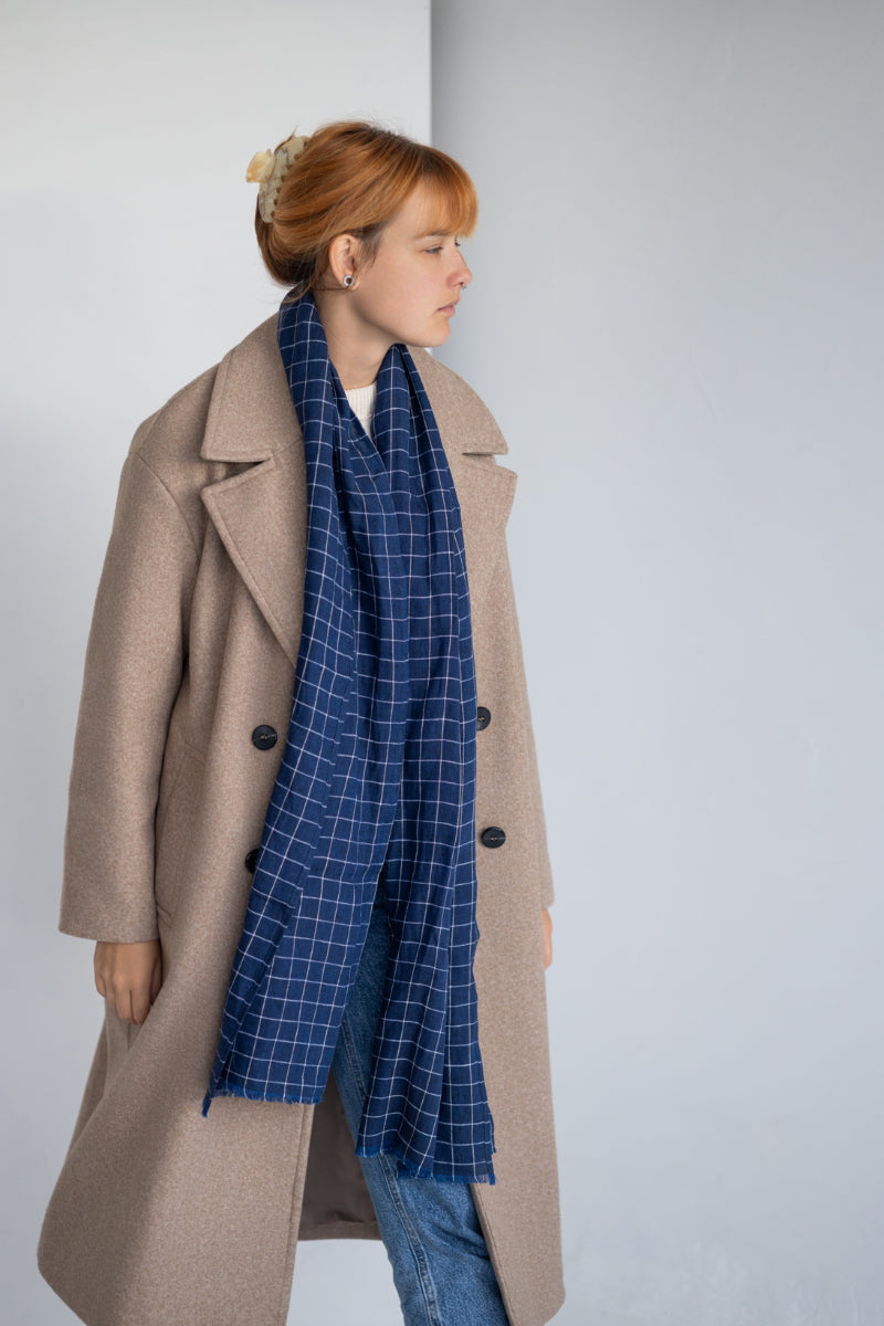 Person wearing a beige coat and blue checkered linen scarf against a white background